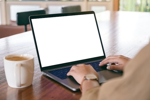 Photo mockup image of a woman using and typing on laptop with blank white screen and coffee cup on wooden table