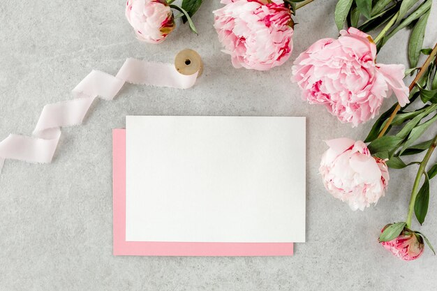 Mockup invitation blank paper greeting card pink envelope and peonies on gray stone table Flower background Flat lay top view