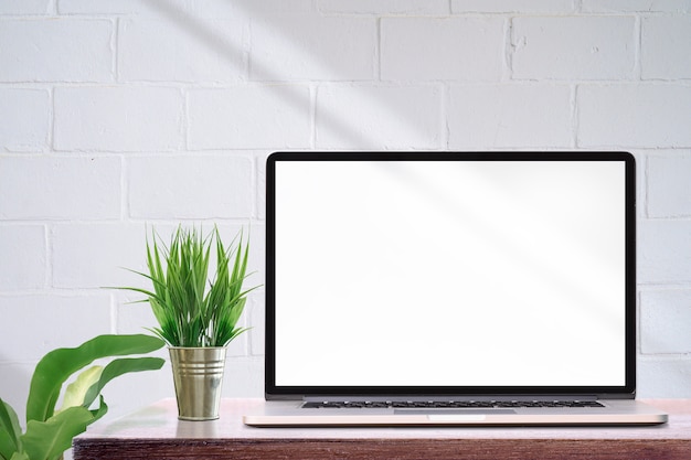 Photo mockup laptop computer with blank screen and houseplant on wooden table, white brick wall and copy space.