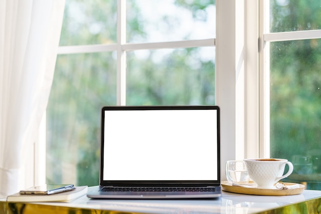 Mockup di computer portatile con schermo vuoto con notebook, tazza di caffè e smartphone sul lato del tavolo la finestra nella caffetteria al bar, schermo bianco