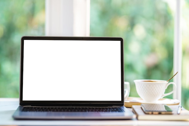 Макет портативного компьютера с пустым экраном с ноутбуком, чашкой кофе и смартфоном на столе у окна в кафе в кафе, белый экран
