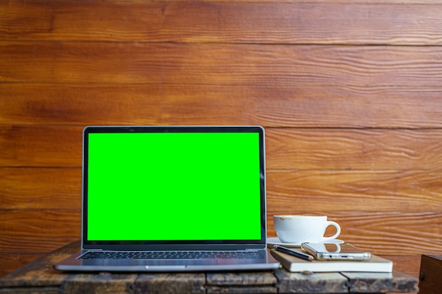 Mockup di computer portatile con schermo vuoto con notebook, tazza di caffè e smartphone sul tavolo dello sfondo della parete in legno al caffè, schermo verde