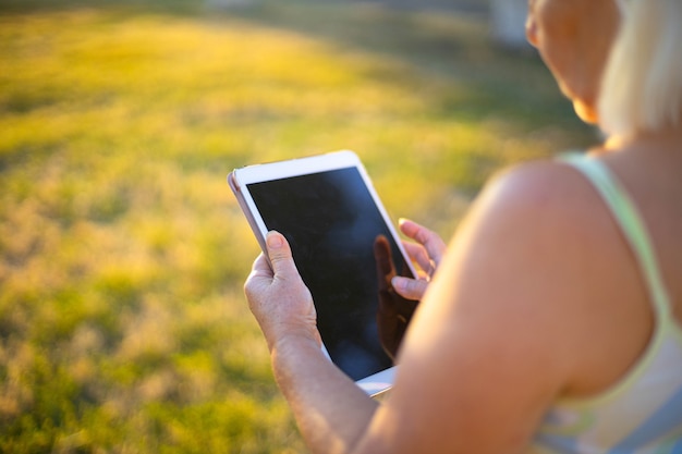 緑の草の上に空白の白い画面でタブレットを座って保持しているモックアップノートパソコンの若い女性