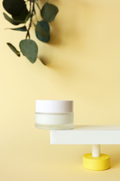 Mockup moisturizer cream in jar standing on abstract pedestal on pastel yellow background with copy space and defocused eucalyptus leaves, front view.