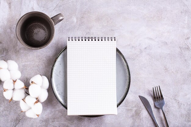 Mockup of a notebook on a plate an empty cup and a branch of cotton on a gray background top view