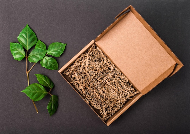 Foto mockup open kartonnen doos met papieren vulling op zwarte achtergrond met groene bladeren bovenkant