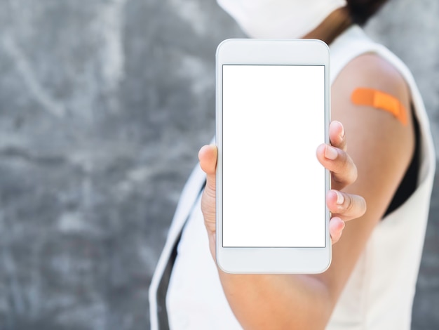 Foto telefono mockup, schermo bianco vuoto sullo smartphone che tiene e mostra la donna vaccinata che indossa un blazer bianco senza maniche, una maschera facciale e un cerotto sulla spalla con spazio per le copie.