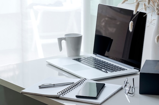 Mockup smartphone on open notebook with laptop, glasses,box and cup on white wooden table.