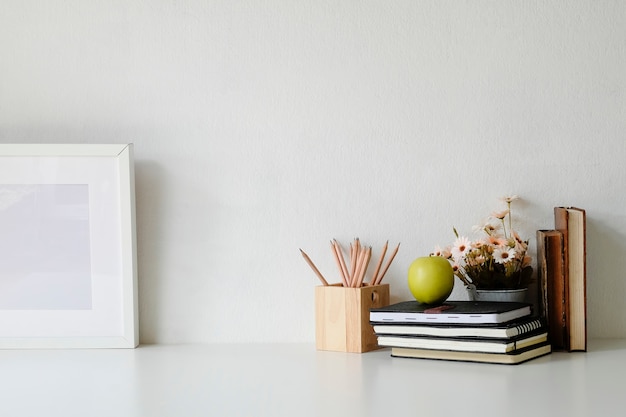 Mockup table with flower, photo frame, books, green apple and jar of pencil on white table.