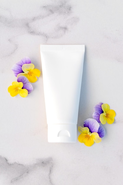 Mockup of unbranded white squeeze bottle tube and small pansy flowers on marble table Top view Natural organic spa cosmetics concept Blank for branding and label