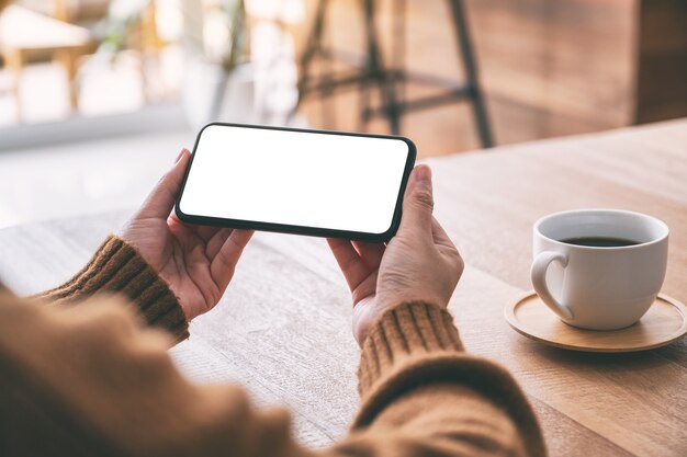 Mockupbeeld van de hand van de vrouw met zwarte mobiele telefoon met leeg scherm horizontaal met koffiekopje op houten tafel