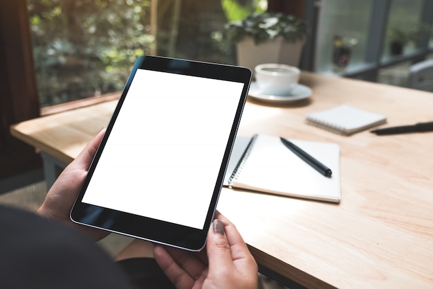 Mockupbeeld van de handen van een vrouw die zwarte tabletpc met wit leeg scherm met notitieboekje en koffiekop op lijst houden