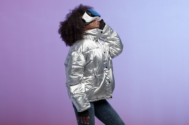 Mod curly brown-haired girl dressed in a silver-colored jacket uses the virtual reality glasses in the studio on neon background .