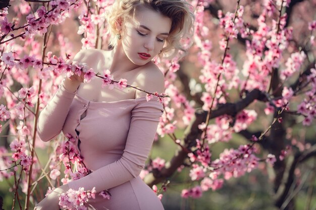 Mode buiten foto van prachtige jonge vrouw in elegante jurk poseren in tuin met bloesem perzikbomen. Blond in bloeiende tuinen