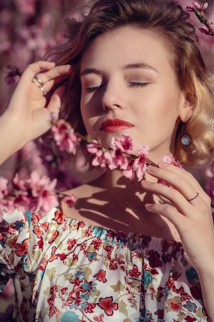 Mode buiten foto van prachtige jonge vrouw in elegante jurk poseren in tuin met bloesem perzikbomen. Blond in bloeiende tuinen