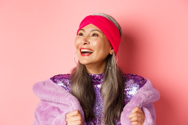 Mode. Close-up van een gelukkige Aziatische senior vrouw die lacht, naar links kijkt met een vrolijk gezicht, staande in een paarse namaakbontjas, roze achtergrond.