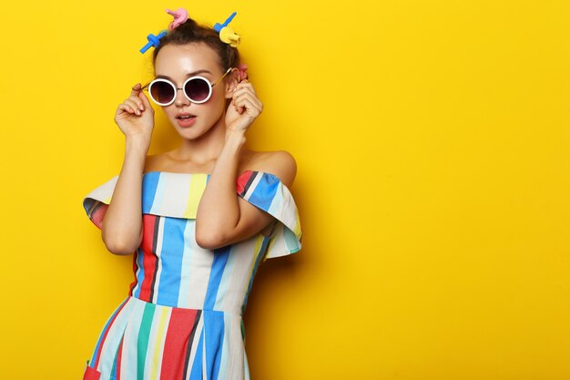 Mode cool meisje poseren in zonnebril op gele achtergrond. Jonge hipster vrouw, krulspelden in haar haar