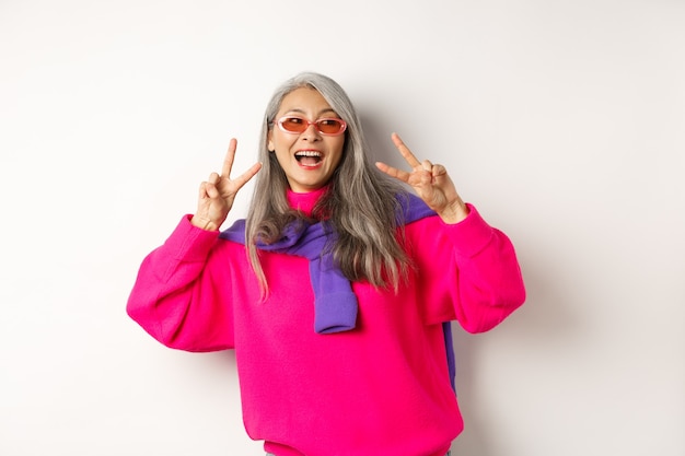 Mode en beauty concept. Afbeelding van een stijlvolle Aziatische senior vrouw met een zonnebril die lacht, vredestekens toont en er gelukkig uitziet, staande op een witte achtergrond