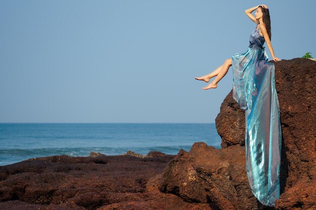 Mode foto van glamoureuze brunette vrouw in een lange jurk met een trein blauwe kameleon kleur nimf op de oceaan met rotsen en golven.