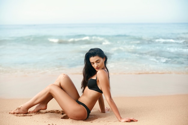 Mode foto van mooie vrouw in zwarte bikini zittend op het zand van het strand