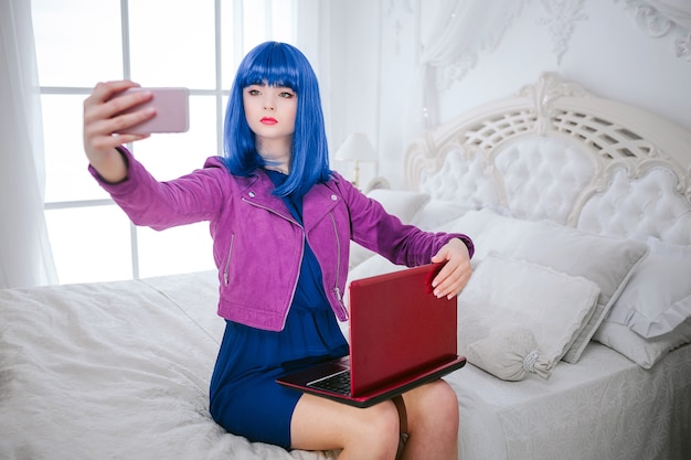 Mode gek. Glamour emotieloos synthetisch meisje, neppop met blauw haar houdt computer vast en doet selfie zittend op het bed.