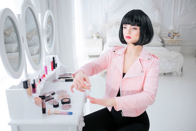 Mode gek. Glamour synthetisch meisje, neppop met lege look en kort zwart haar schildert haar nagels terwijl ze in de buurt van de spiegel zit. Stijlvolle vrouw in roze jas in de slaapkamer. Mode en beauty