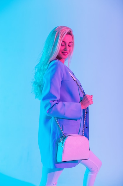 Mode gekleurd portret van mooie jonge lachende vrouw in modieus elegant pak met blauwe blazer en stijlvolle witte lederen handtas op neon roze licht in studio