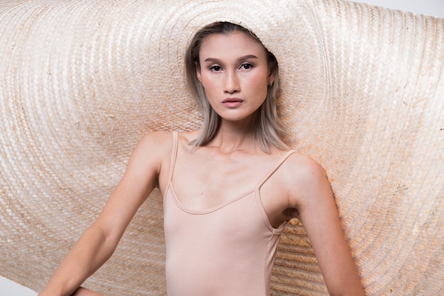 Foto mode jonge dunne aziatische vrouw tan huid ogen mooie make-up mode roze jurk poseren aantrekkelijke glamour look zeer grote strohoed. studio verlichting gebroken witte achtergrond, zomer strand hete zon