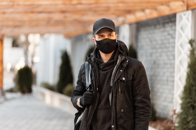 Mode jonge knappe man met een zwart beschermend masker in modieuze kleding met een winterjas, pullover en stijlvolle zwarte pet met rugzak op straat. Coronavirus-concept