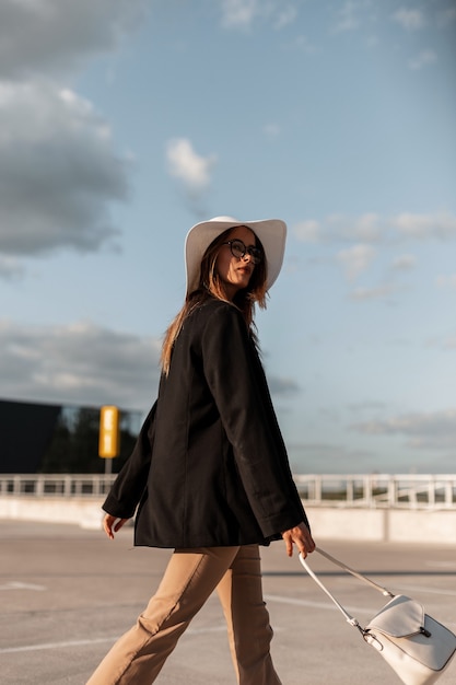 Mode jonge vrouw in zwang zwarte blazer in stijlvolle strohoed in elegante broek met tas loopt op parkeerplaats in de stad in zonnige dag. Nieuwe trendy casual outfit voor dames.