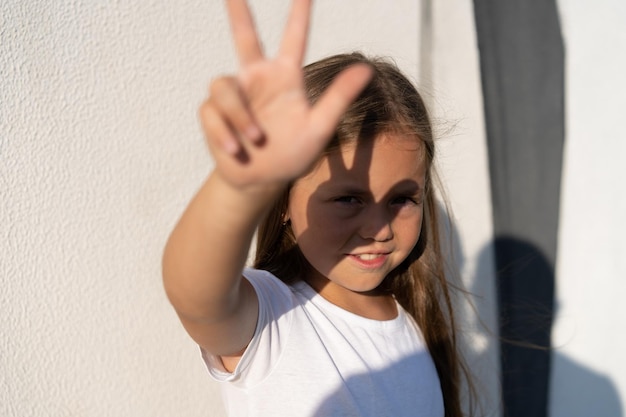 Mode kid concept portret van een meisje dat haar gezicht bedekt tegen de felle zon terwijl ze tegen de witte muur staat