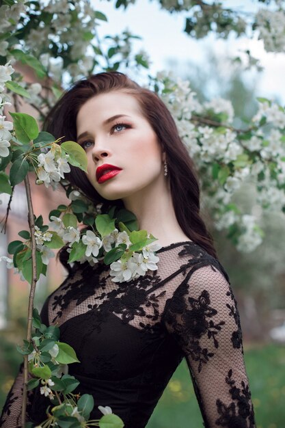 Mode kunst Vrouw in de zomer in de buurt van bloeiende appelboom, felle lippenstift. Lente zomer stemming, mysterieus romantisch beeld van meisje in een zwarte jurk boom bloemen. Vrouw dromen