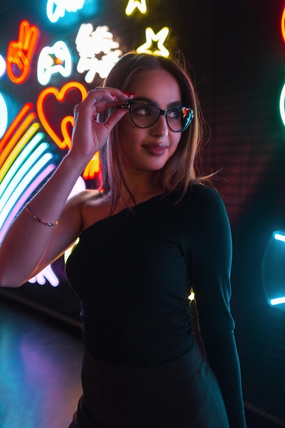 Mode mooie vrouw in zwarte kleding draagt een stijlvolle bril op een donkere achtergrond met kleurrijke neonlichtreclames Urban night female portrait of beauty girl