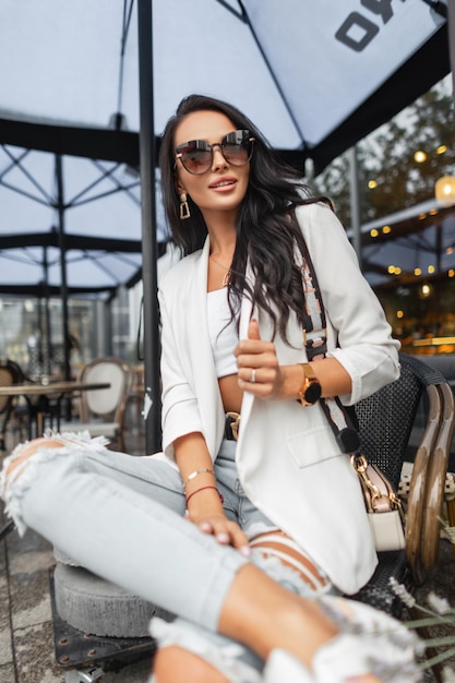 Mode mooie zakenvrouw met stijlvolle zonnebril in trendy casual witte outfit met witte blazer top en gescheurde spijkerbroek met modetas zit op stoel en rust in café op straat Stedelijke dame