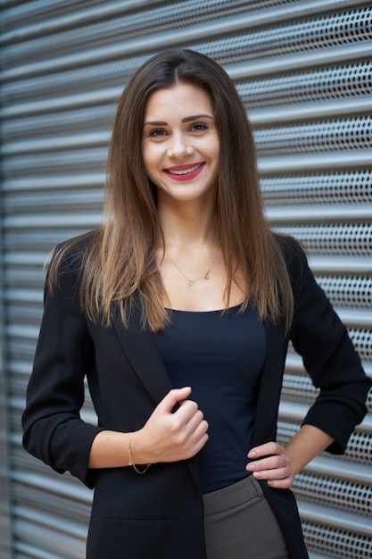 Mode. Portret van aantrekkelijke gelukkig jong meisje met charmante glimlach dragen van stijlvolle zwarte jas, bruine broek en elegante zwarte schoenen in de buurt
