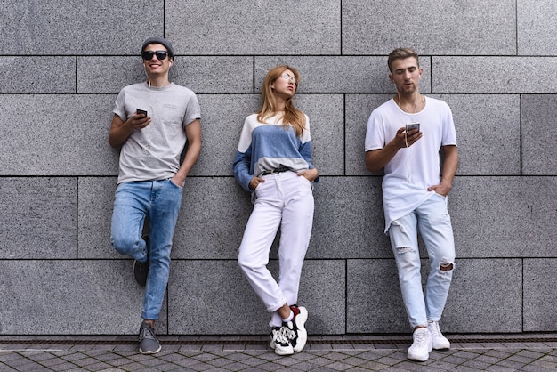 Mode portret van drie beste vrienden poseren op straat, stijlvolle outfit en jeans dragen tegen grijze muur.