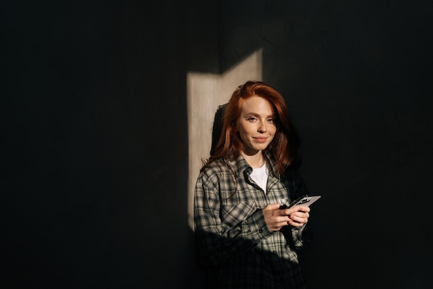 Mode portret van een mooie roodharige jonge vrouw met een mobiele telefoon op schaduw achtergrond in