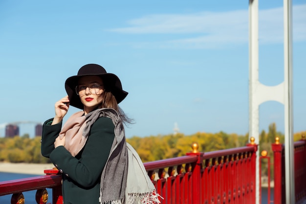 Mode portret van elegante jonge model poseren op de brug. Vrouw met modieuze hoed, sjaal en bril