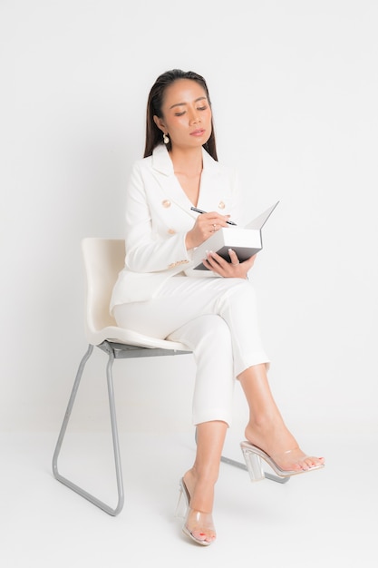 Mode-stijl catalogus kleding voor zakenvrouw zwart lang haar natuurlijke make-up draag wit pak zittend op een witte stoel, boek en pennen te houden bij studio shoot op witte achtergrond.