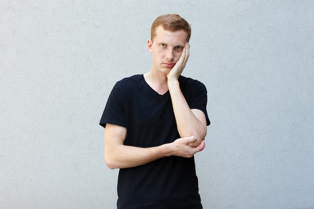 Mode, stijl, emoties en mensen concept - close-up portret van een roodharige van een mooie mannelijke man met sproeten op een grijze achtergrond zwart T-shirt. hoofd vasthouden met hand moe zijn?