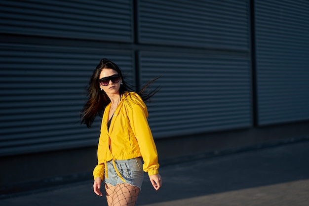 Mode. Stijlvolle hipster brunette meisje gekleed jeugd gele jas, denim shorts en modieuze zonnebril poseren op grijze metalen stedelijke muur