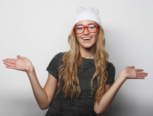 Mode studio portret van mooie jonge hipster blonde vrouw met bril, stijlvolle stedelijke t-shirt en hoed dragen, op witte achtergrond