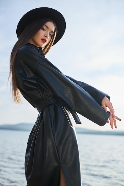Mode vrouw in zwarte ronde hoed en leren regenjas in de buurt van de vijver van het meer. Portretmeisje bij zonsondergang. Mooie make-up en felrode lippenstift op de lippen