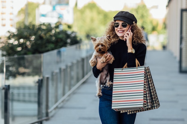 Mode wandeling vrouw met lang krullend haar houdt kleine hond en boodschappentassen Mooi meisje knuffels hondje Glimlachende aantrekkelijke vrouw met Yorkshire terrier Meisje met hond in handen en verkoop concept