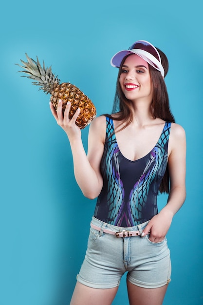 Mode zomer portret mooie vrouw in roze zonneklep en ananas over blauwe achtergrond