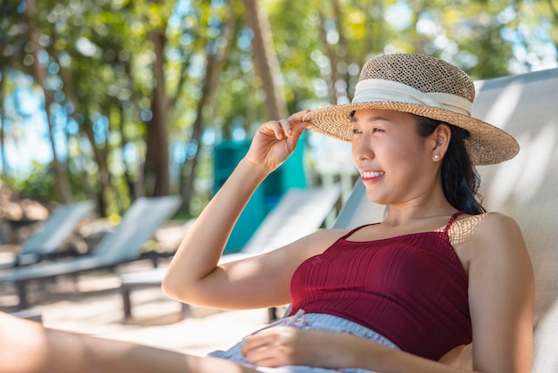 Model Aziatische vrouw met een rood zwempak en een strohoed die op de ligstoel in de zon aan zee ligt