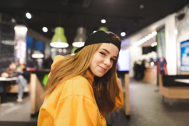 Model in a cap and an orange hoodie poses to the camera indoors