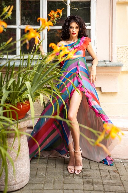 A model in a colorful dress stands in front of a plant with yellow flowers.