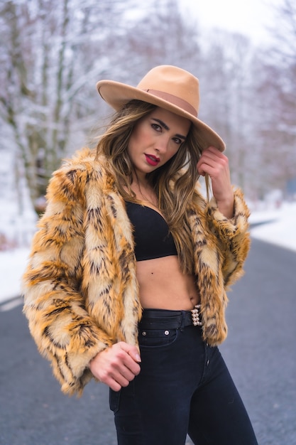 Modello che gode in biancheria intima, un cappello da cowboy e un maglione leopardato in mezzo alla strada con alberi congelati sullo sfondo, stile di vita invernale