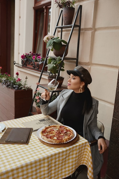 Фото Модель девушки в модной одежде сидит на открытом воздухе за столиком в кафе, пьет вино и ест пиццу